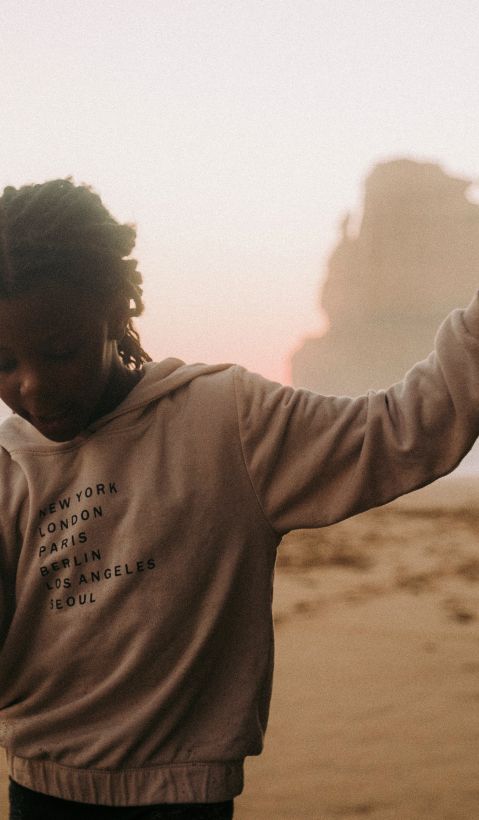 Black girl on the beach with a grey hoodie on, raising her hand joyfully.